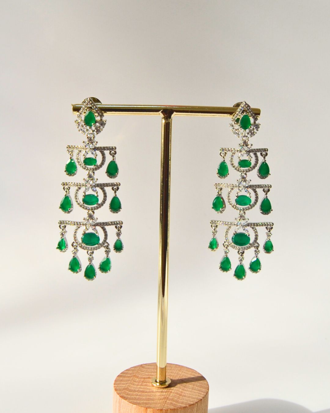 Pair of emerald and diamond chandelier earrings on a gold stand with a wooden base.