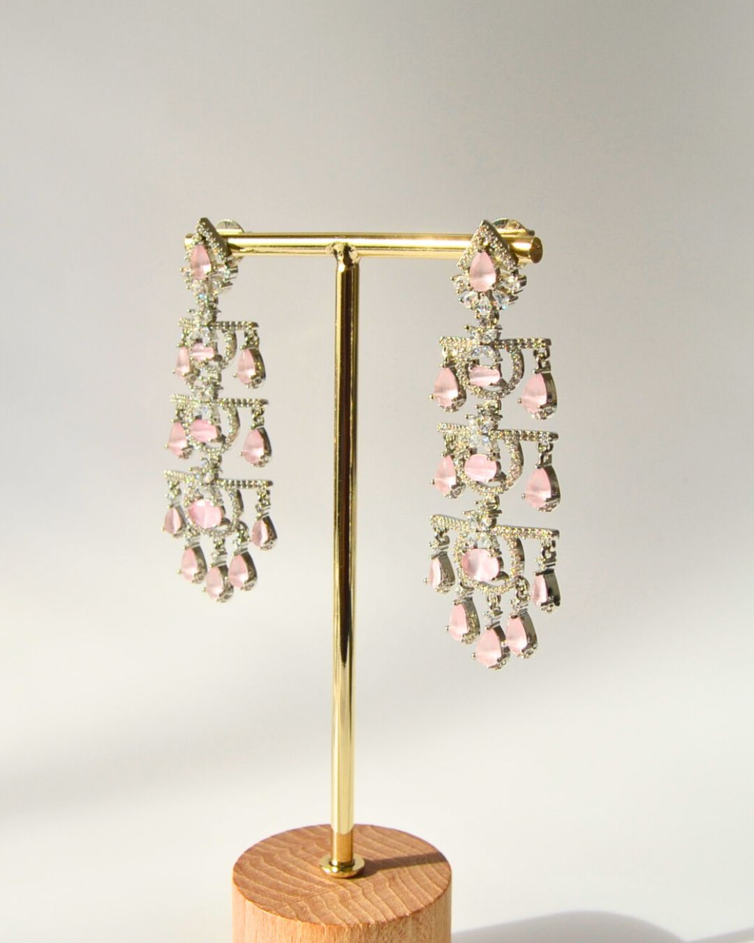 Pair of pink and gold earrings on a gold stand with a wooden base against a light gray background