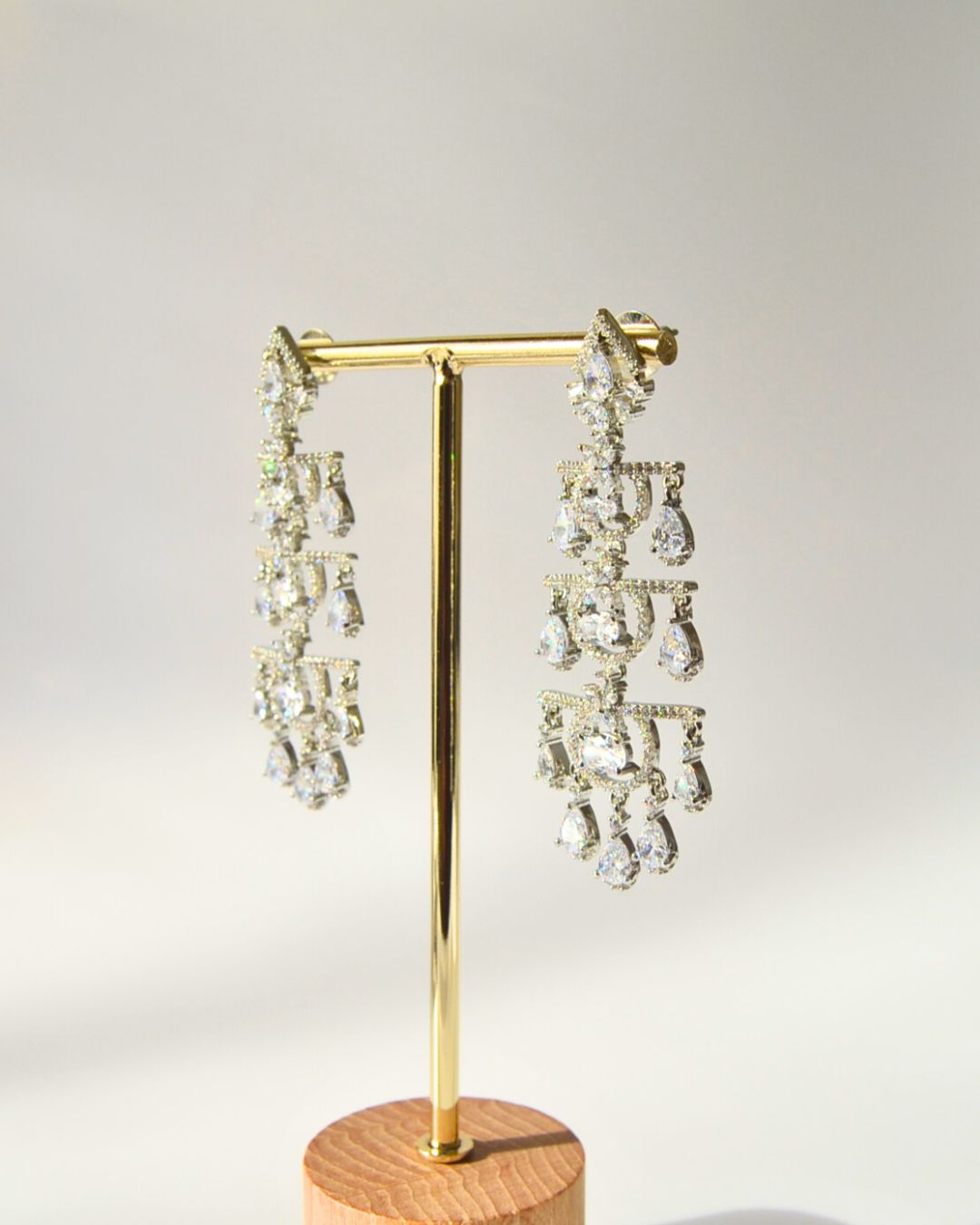 Pair of zircon chandelier earrings on a gold earring stand with a wooden base against a light gray background