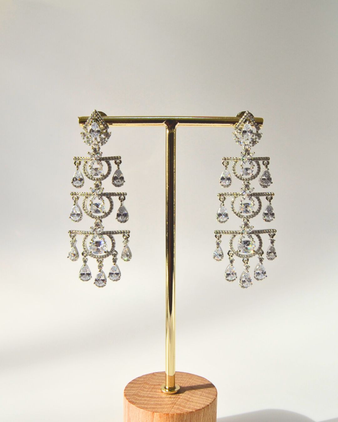 Pair of zircon chandelier earrings on a gold stand with a wooden base against a light gray background
