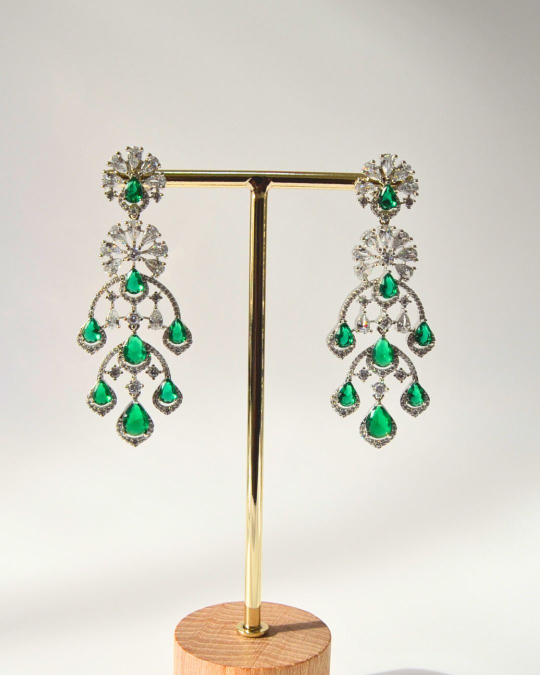Pair of emerald zircon chandelier earrings on a gold stand with a wooden base.